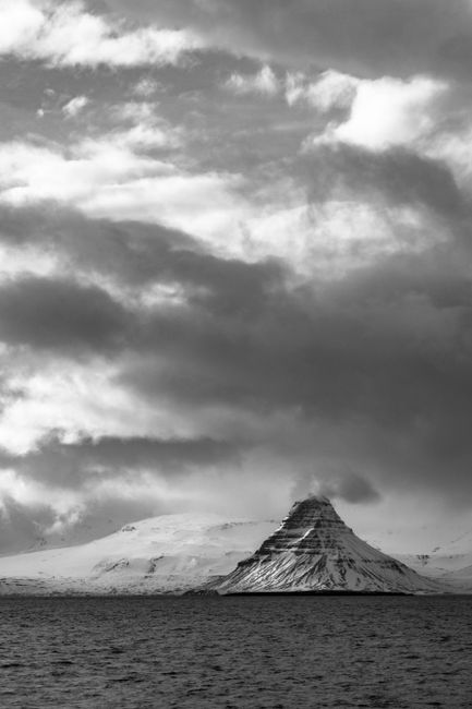 A striking black and white photograph of a snow-covered mountain under a dramatic cloudy sky over the ocean.