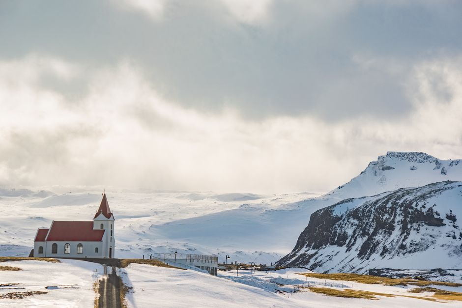 A picturesque church stands amidst a snowy winter landscape with mountains and a cloudy sky.