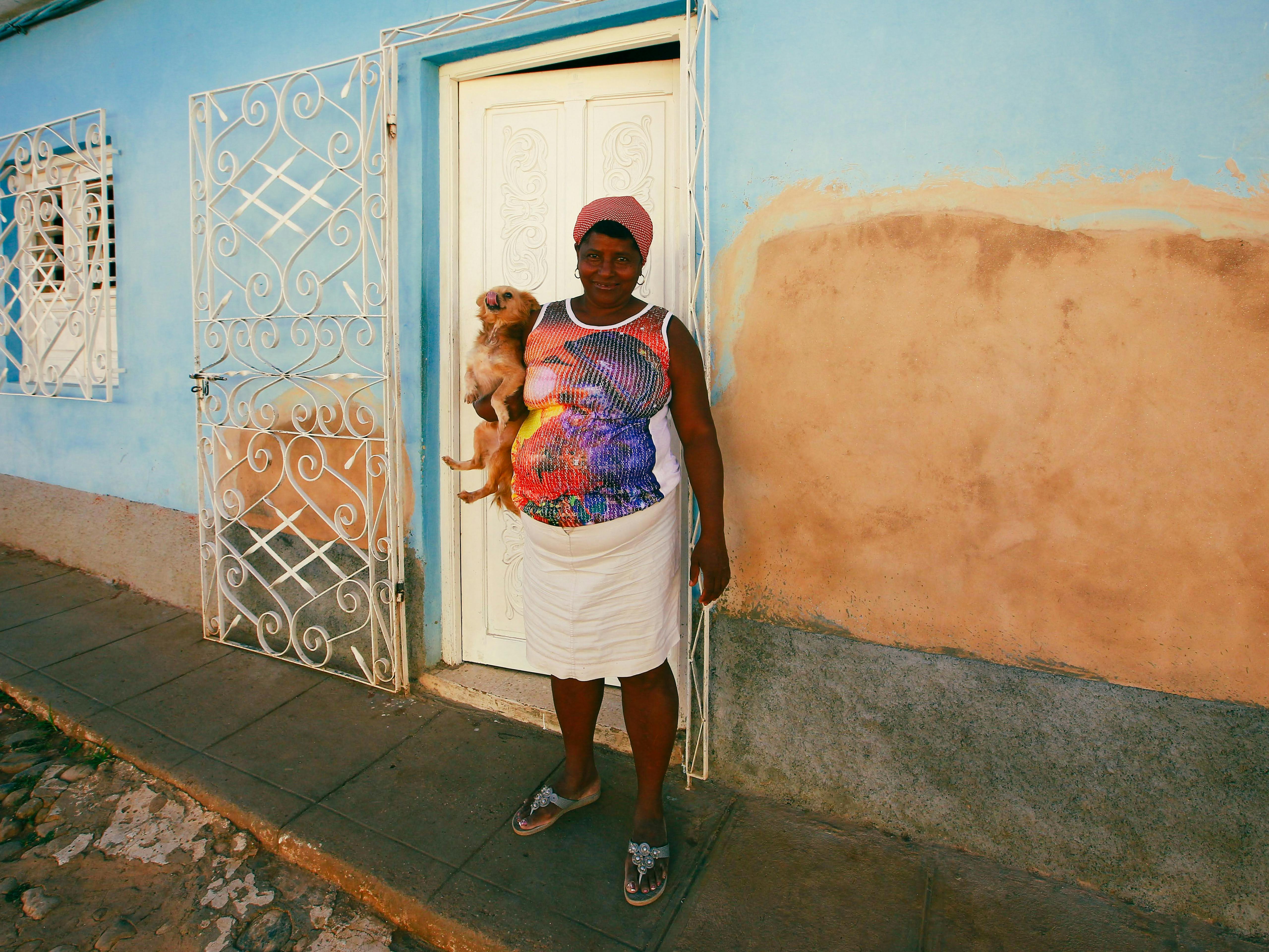 Woman holding dog in front of colorful Mediterranean style house with ornate iron door, displaying cultural architecture.