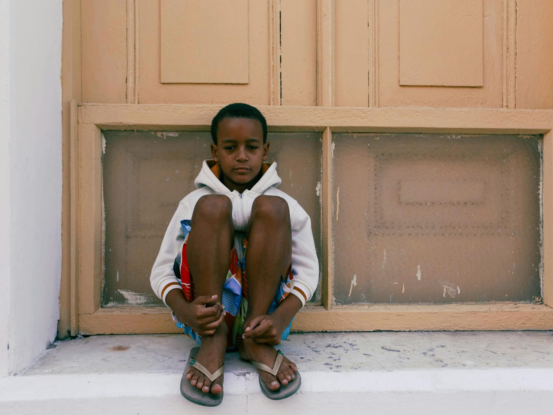 A young boy sits thoughtfully on a doorstep in a street scene. Calm and reflective mood.
