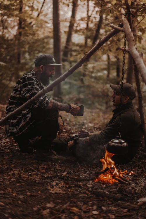 Free Two men enjoying a campfire in an autumn forest, sipping drinks and sharing stories under a canopy of trees. Stock Photo