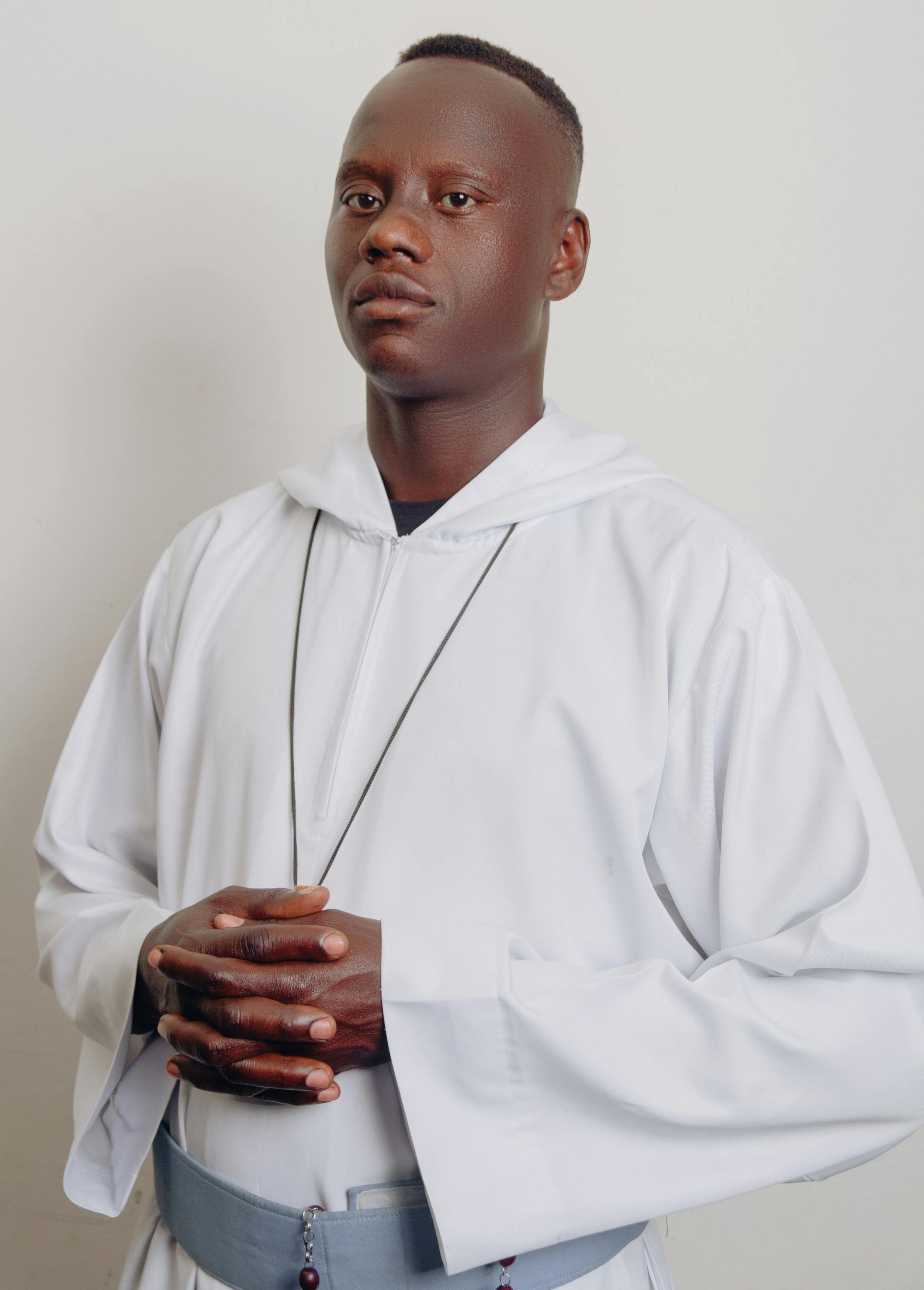 A man in white robes stands with hands together, conveying calm focus indoors.