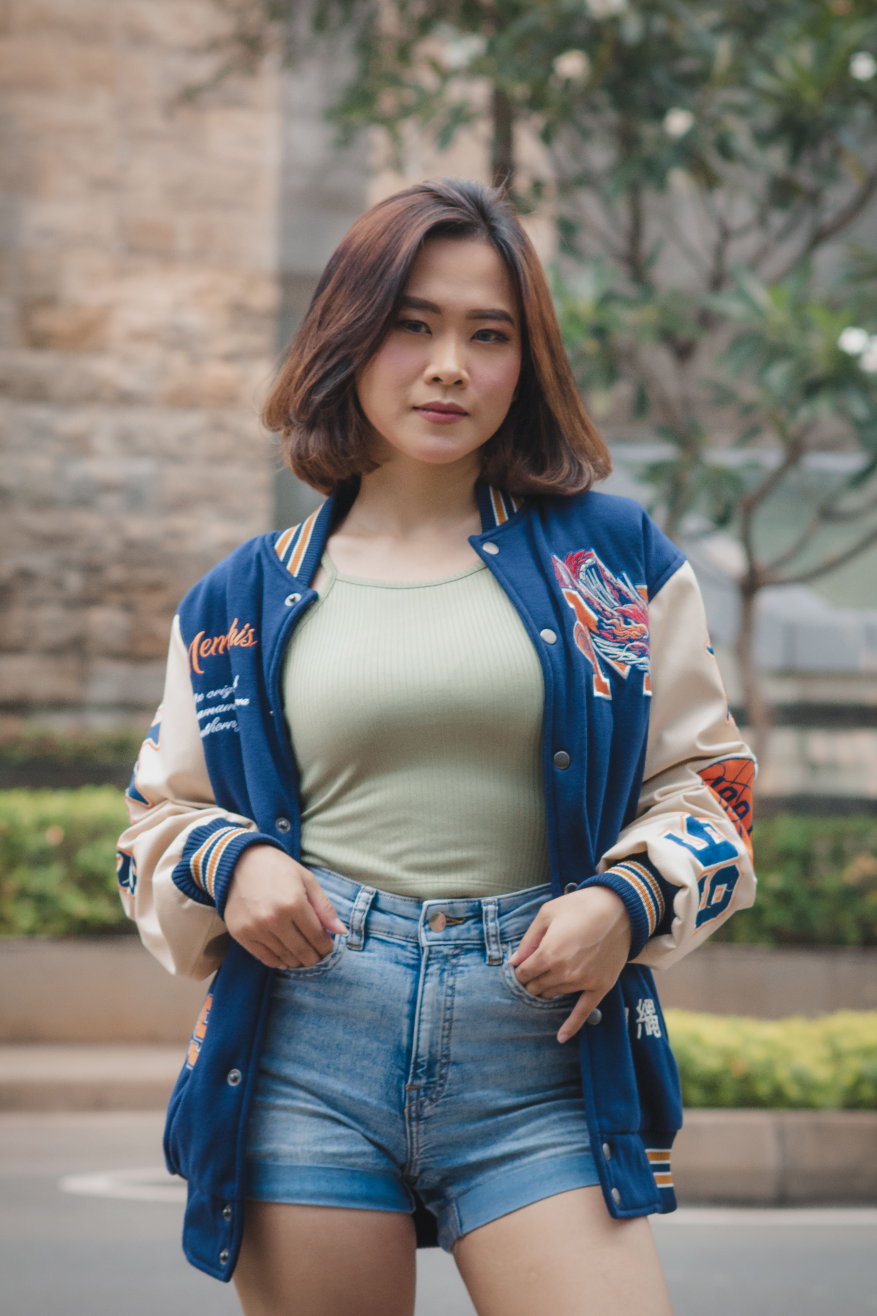 Trendy teen in denim shorts and varsity jacket posing outdoors, exuding confidence.