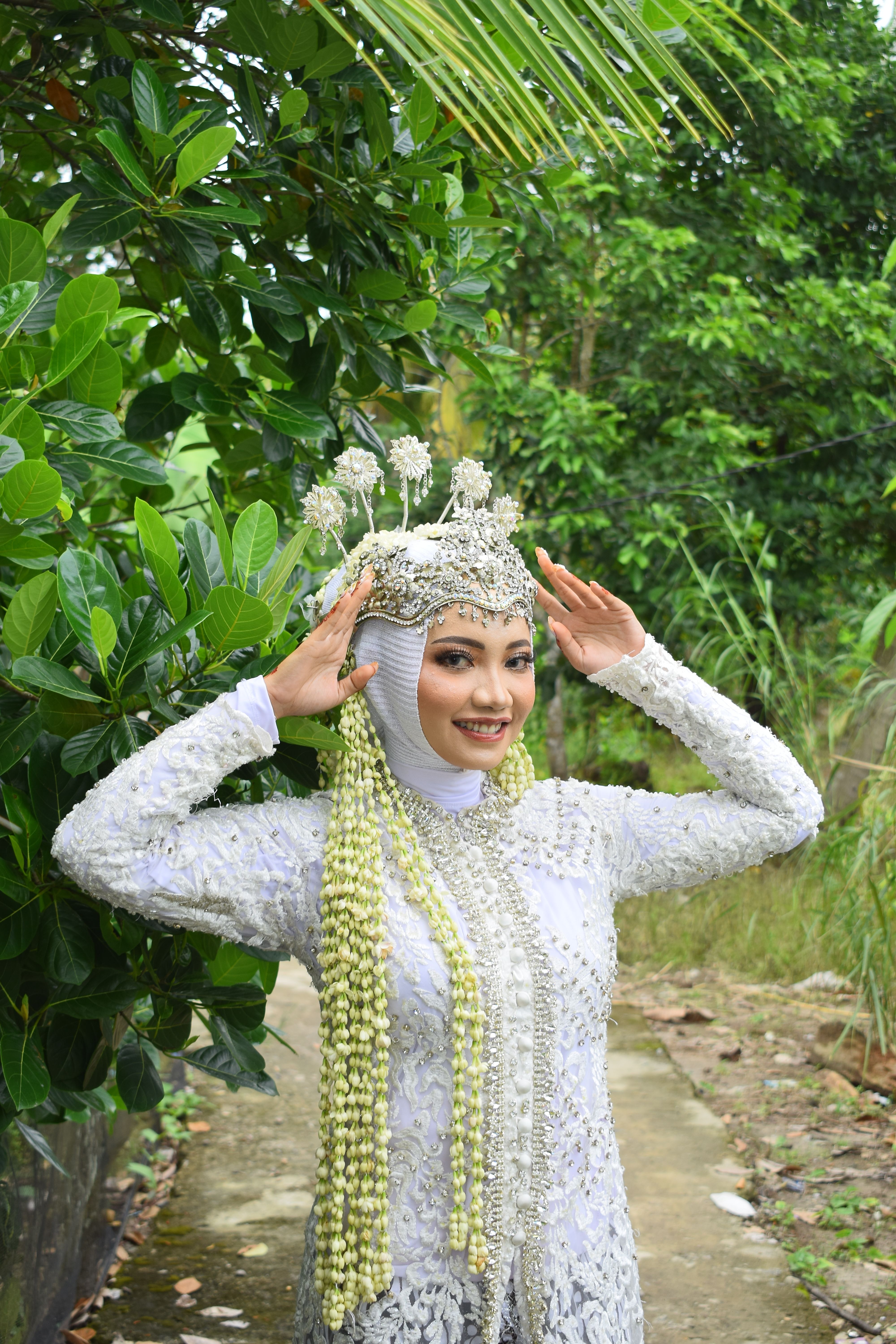Smiling bride in intricate traditional attire posing outdoors amidst lush greenery.