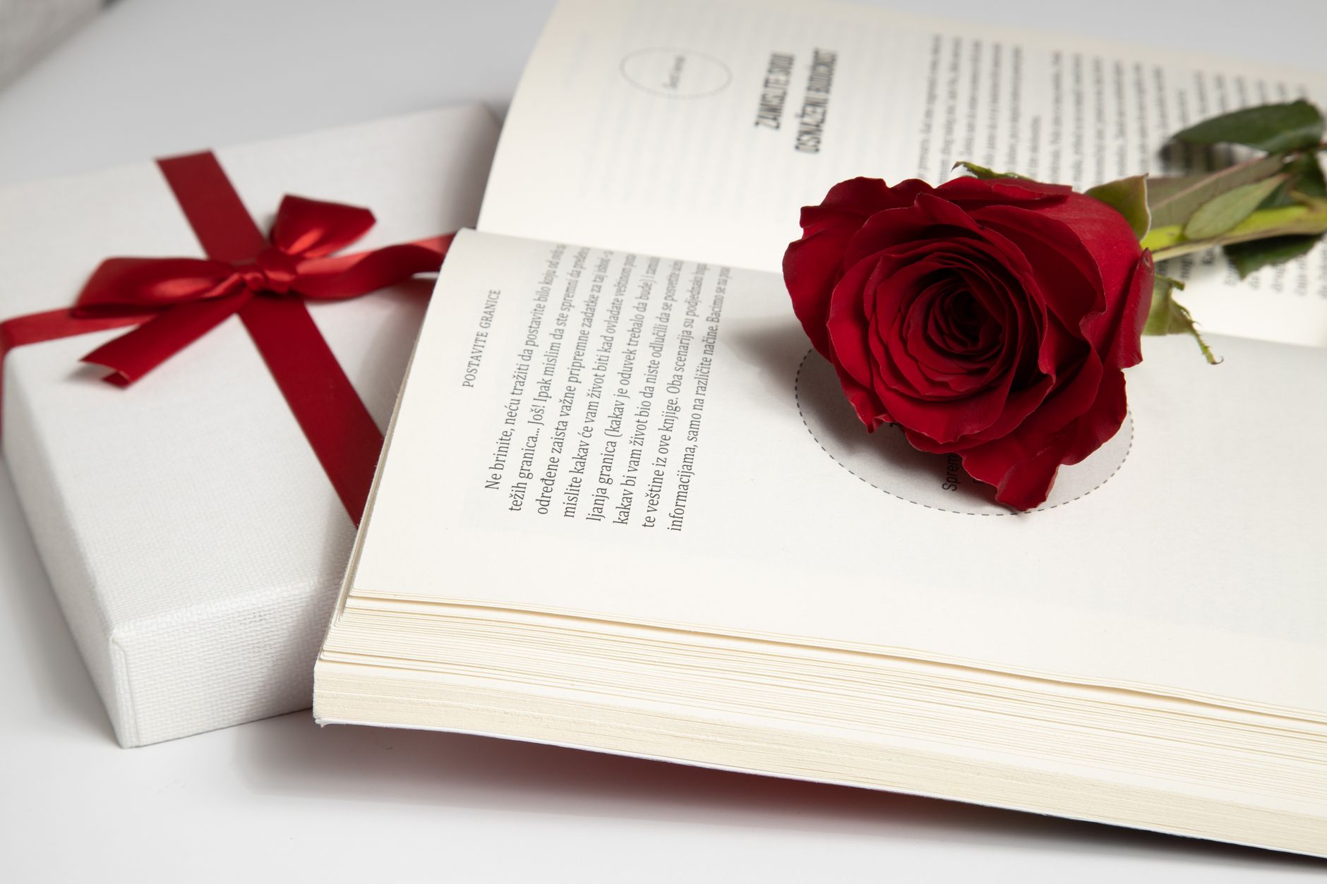 A red rose placed on an open book beside a gift box wrapped in ribbon, symbolizing romance and love.