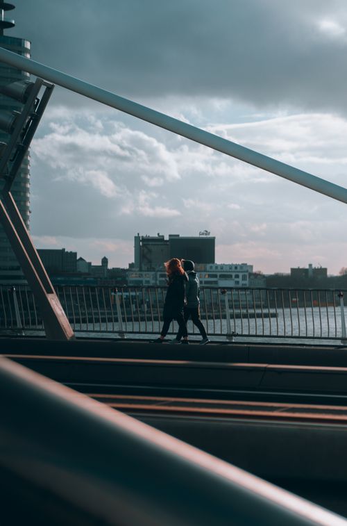 Free Two people walk on a modern bridge overlooking city buildings under a cloudy blue sky. Stock Photo