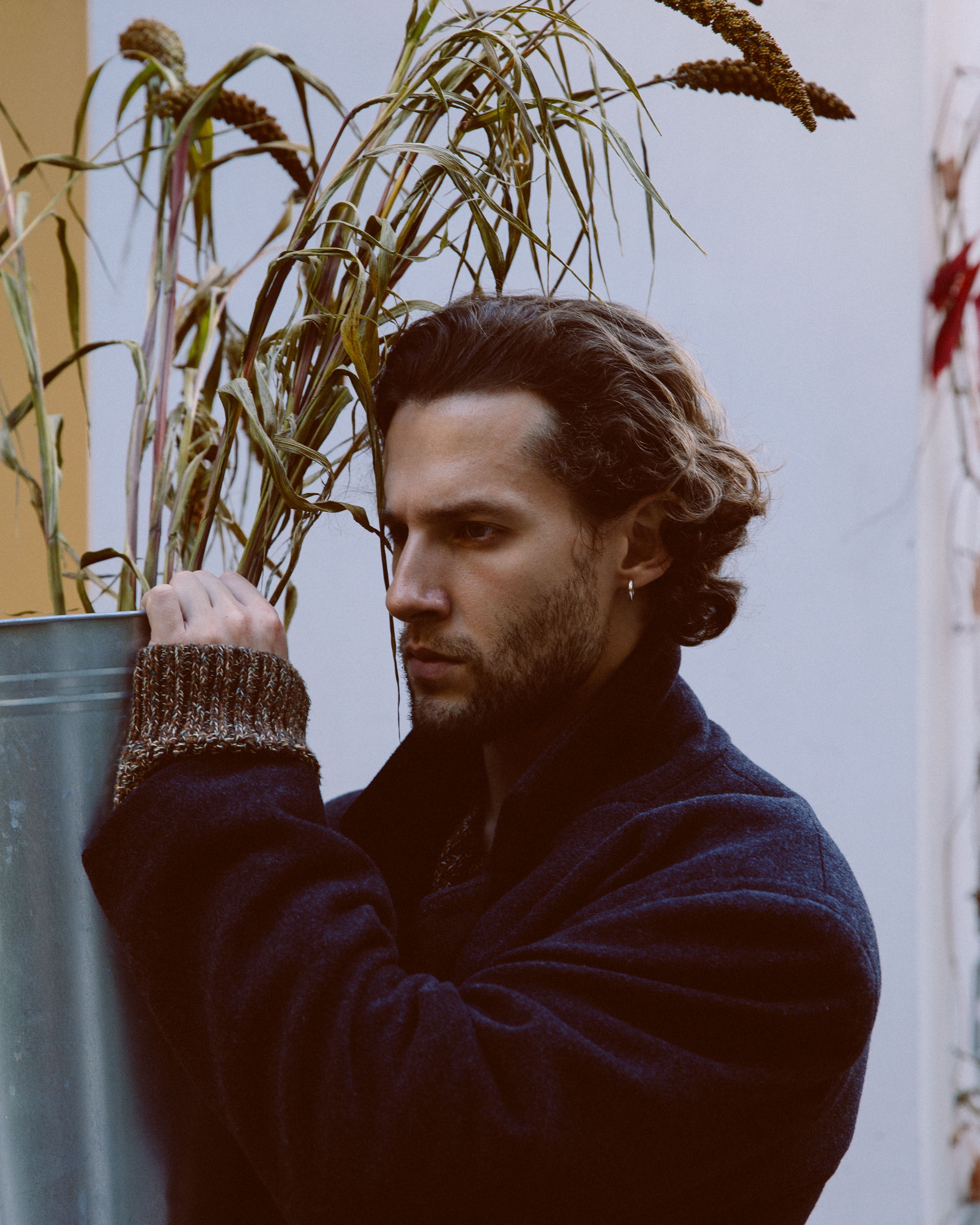 A contemplative man in a dark coat posing with an ornamental plant against a subtle indoor background.