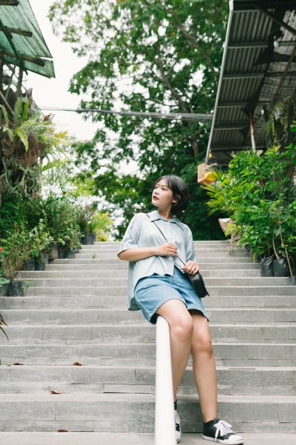 Casually dressed woman sitting on stairs with greenery around during the day.