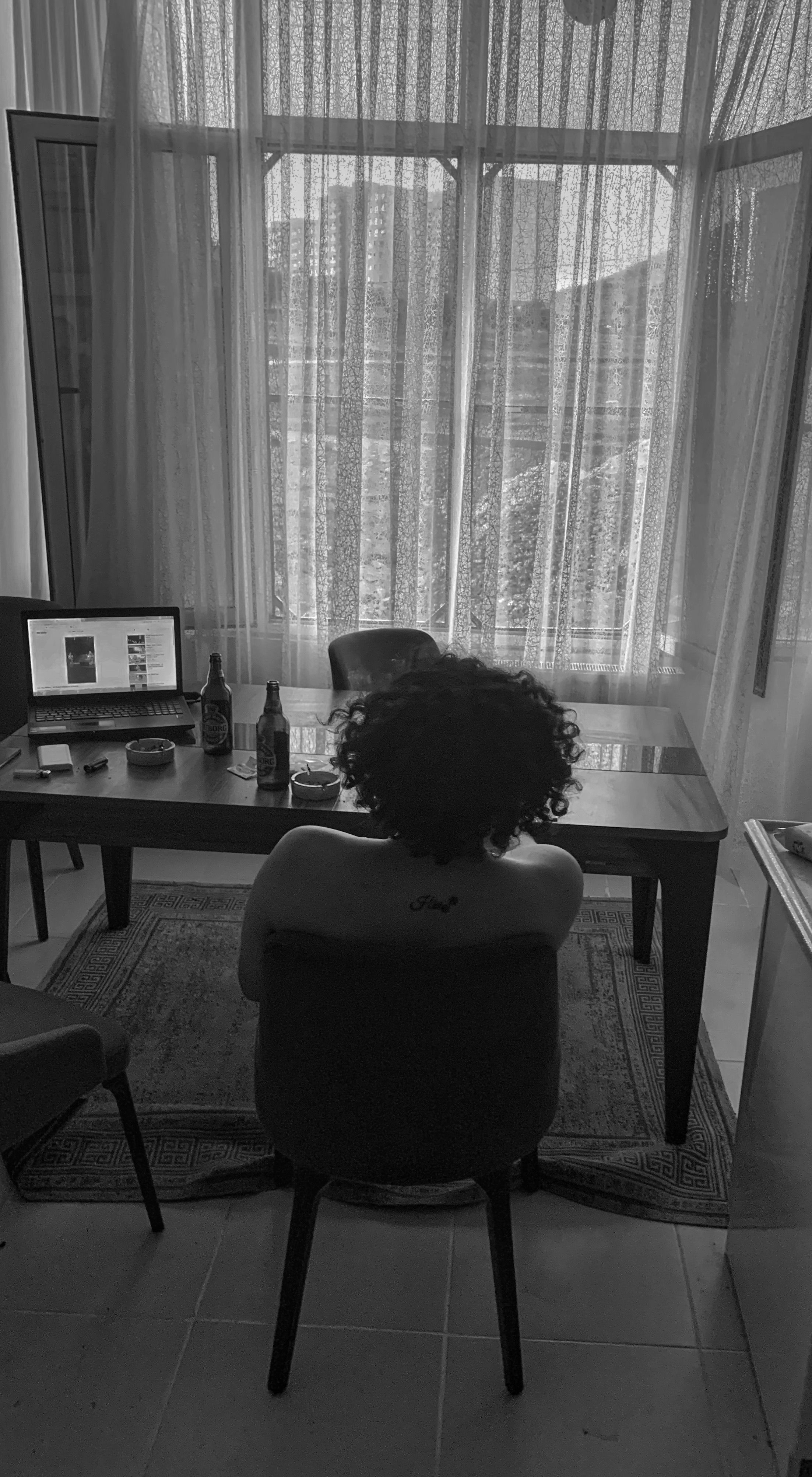 Monochrome image of a person seated, gazing out through a large window, in a serene setting.