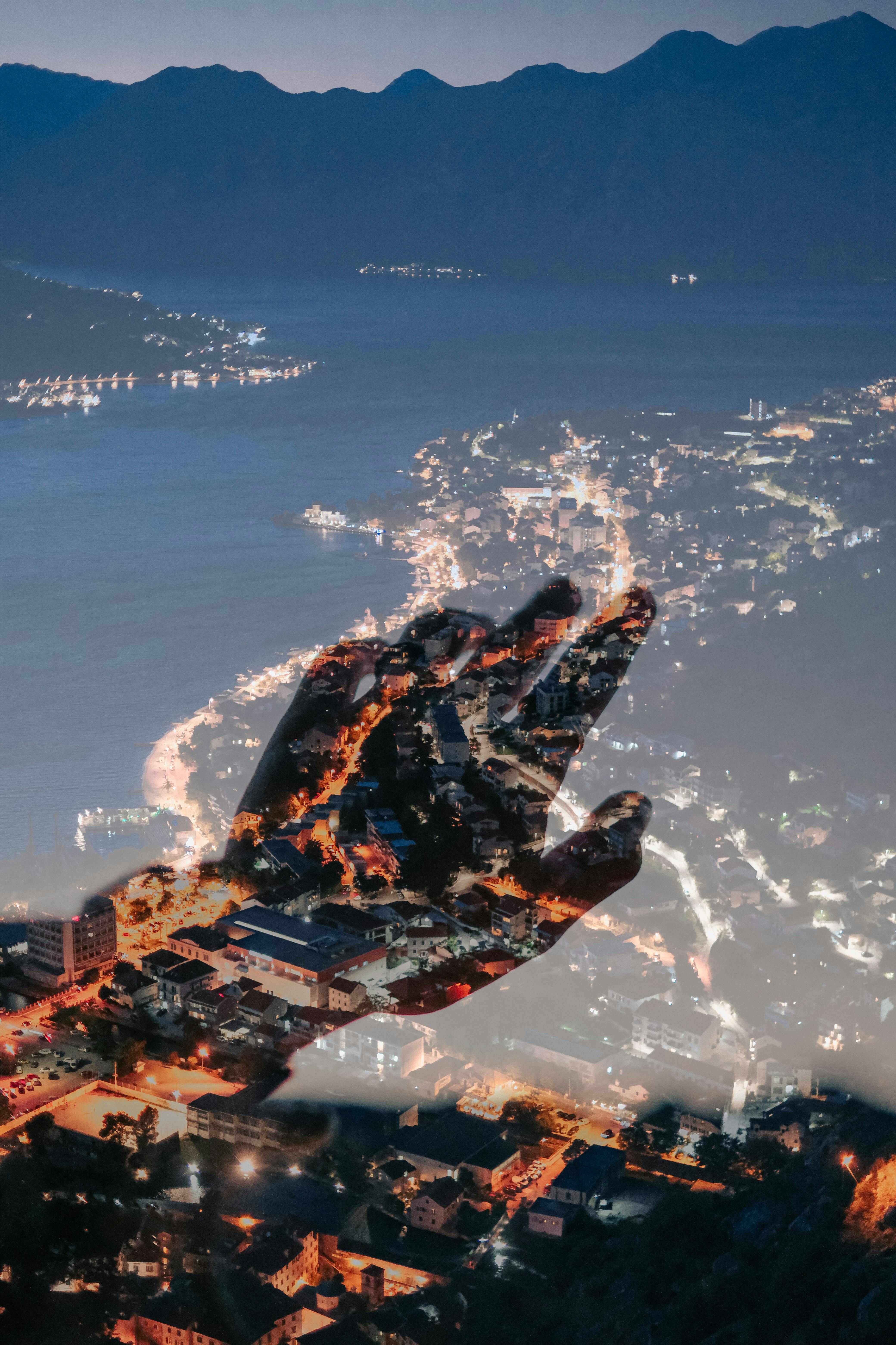 Intriguing double exposure of a cityscape with a hand silhouette over illuminated coastline at night.