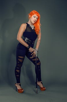 A fashionable woman with orange hair poses confidently in a studio setting wearing stylish black attire.
