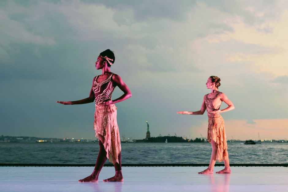 Two graceful dancers perform outdoors near the Statue of Liberty at sunset, showcasing elegance and poise.
