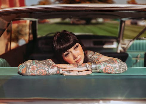 A tattooed woman with bangs leans on the seat inside a vintage car, basking in sunlight.