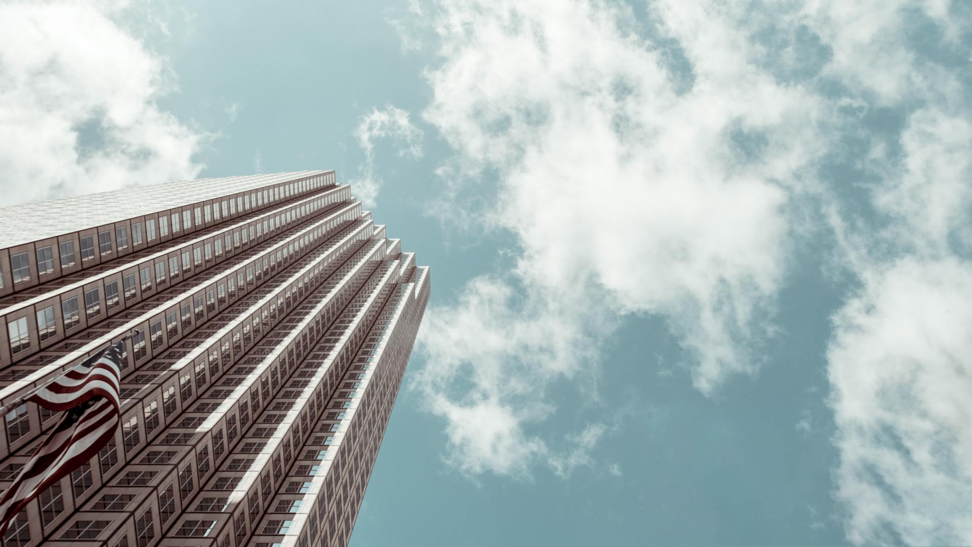 Low-angle view of a modern skyscraper with American flag and cloudy sky backdrop.