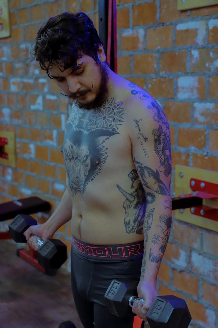 Shirtless Young Man With Tattoos Holding Dumbbells In Gym