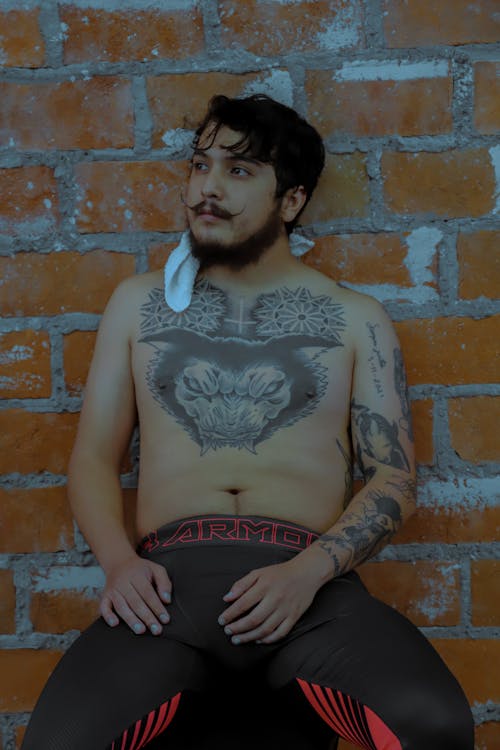 A Shirtless Tattooed Man Leaning on a Brick Wall