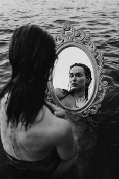 Black and white photo of a woman holding a mirror with her reflection in the water.