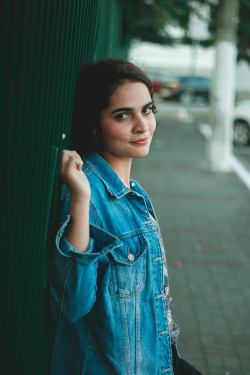 Free Charming young woman leans against a fence, wearing a denim jacket and smiling. Stock Photo