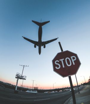 A dramatic shot capturing an airplane flying over a road with a stop sign, silhouetted against the sunset sky.