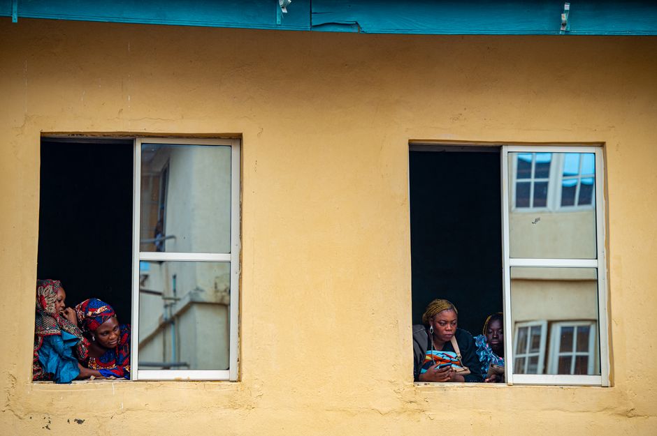 Four African women peering through windows in a yellow building facade.