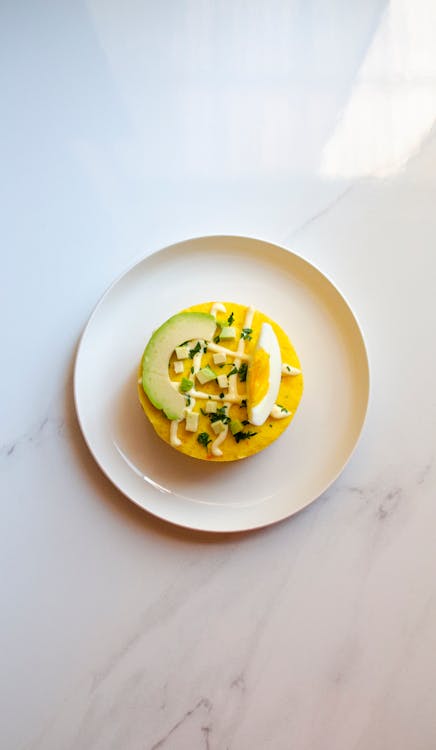 A white plate placed on a white marble surface that contains a causa with a topping of avocado.