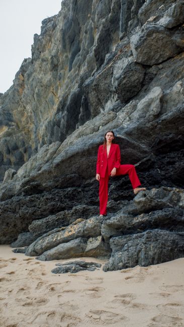 Woman in a vibrant red suit posing confidently on a rocky beach setting. Fashion meets nature.