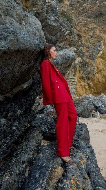 Stylish woman in red suit leaning against rocky seaside, exuding elegance and confidence.