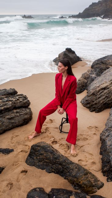 Fashion model in red suit poses barefoot on a rocky seashore, blending style with nature.