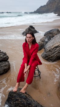A woman in a red suit sits barefoot on a rocky beach, highlighting a serene coastal scene.