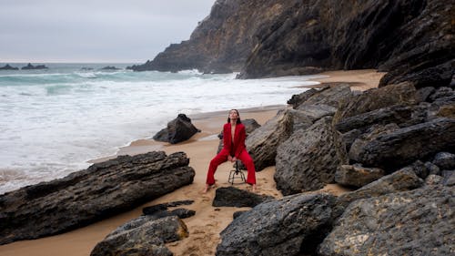 Woman Posing Barefoot in Red Suit Sitting on a Stool on Seashore
