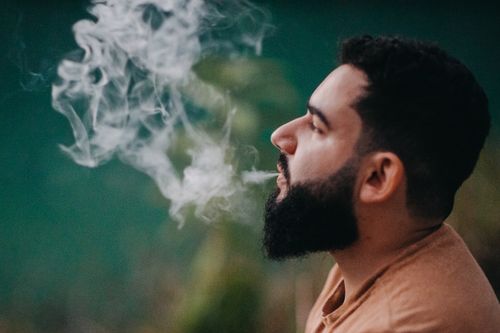 Free Side profile of a bearded man exhaling smoke outdoors with a blurred background. Stock Photo