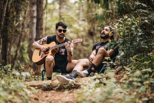 Two men playing guitar and singing in a forest, enjoying music outdoors.