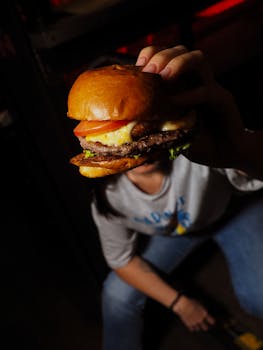 A hand holding a freshly made beef burger with cheese, lettuce, and tomato in a dark setting.