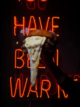 A delicious cheesy burger is held up against a vivid neon sign backdrop, creating a striking food photography composition.