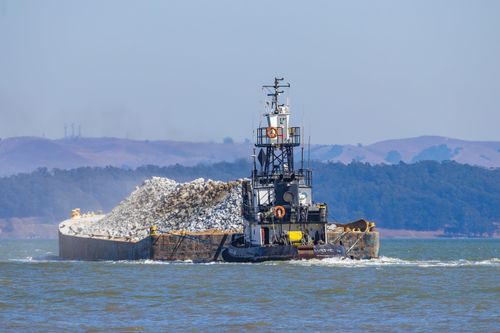Free Tugboat hauling a barge filled with rocks across a scenic sea against a mountainous backdrop. Stock Photo