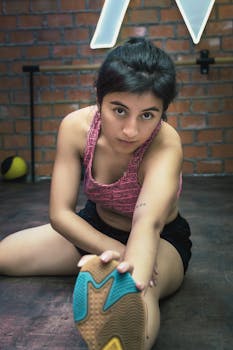 A young woman stretching in sports attire inside a gym with brick walls.