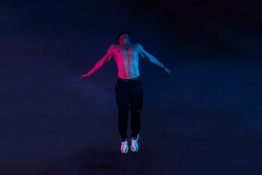 A shirtless man striking a powerful pose in mid-air against a dramatic dark background.