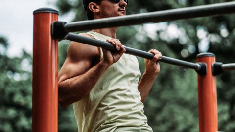 Man in white tank top exercising outdoors on metal bars.