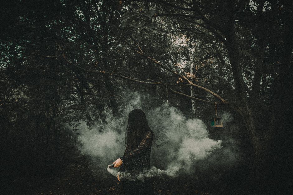An enigmatic woman in black surrounded by smoke in a dark forest.