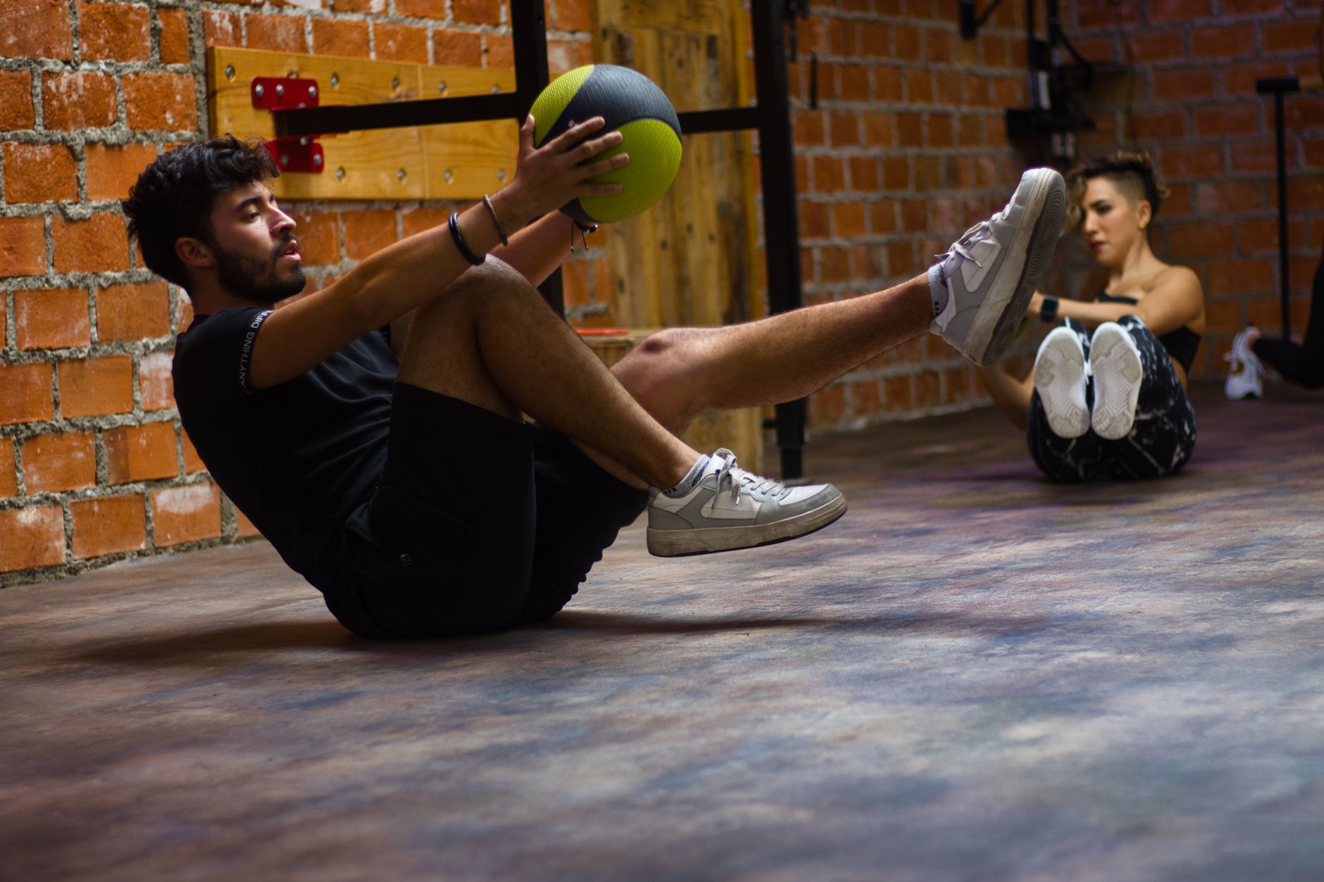 A man and woman perform core exercises with a medicine ball in a brick wall gym.