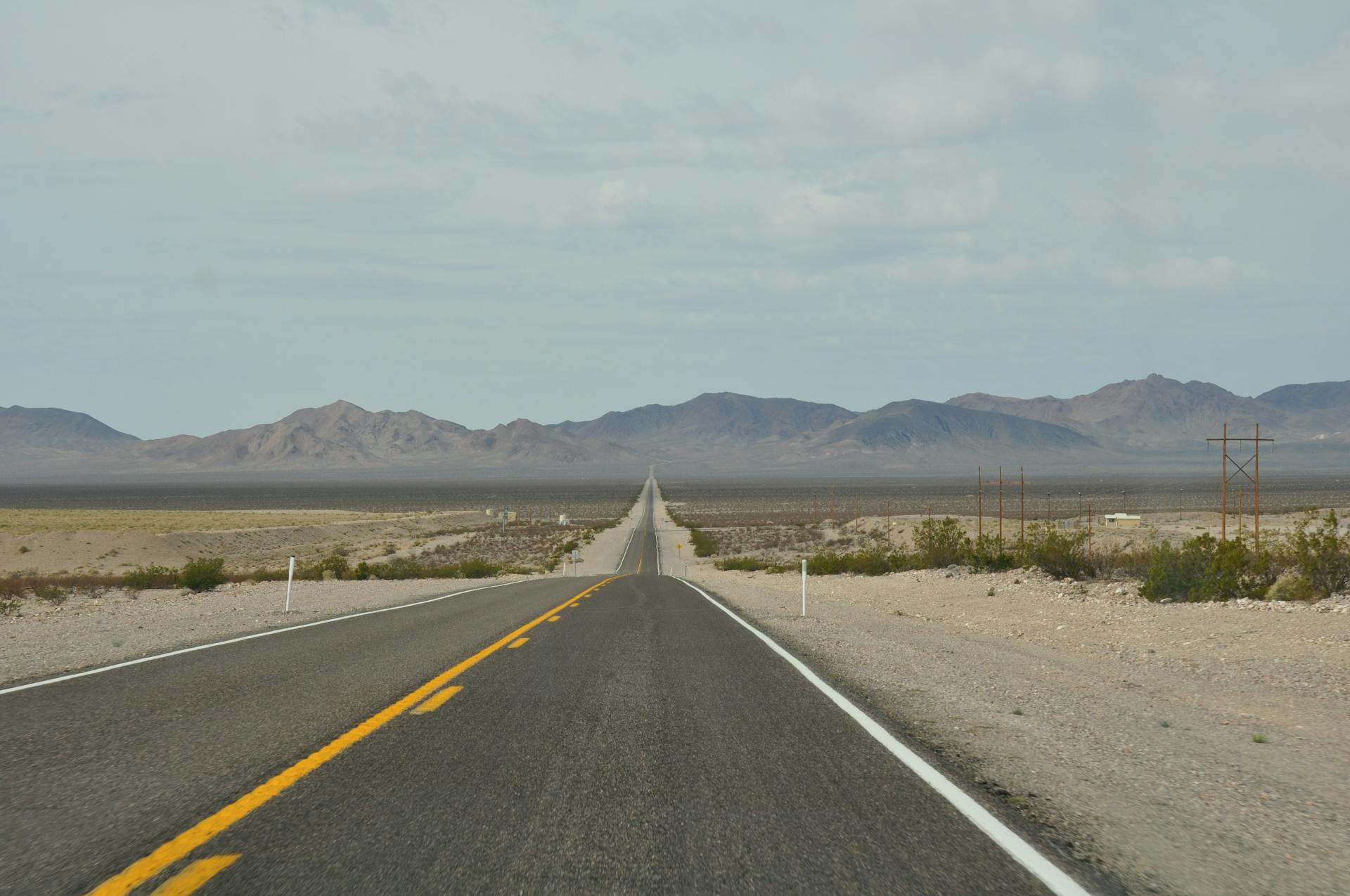 Long straight road through arid desert landscape towards distant mountain range under cloudy sky.