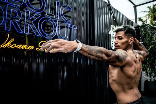 Fit muscular man stretching topless beside neon gym signage, outdoors.