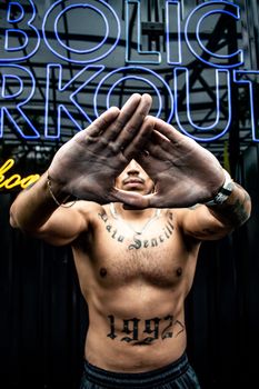 Muscular tattooed man posing shirtless with hands covering face in gym.