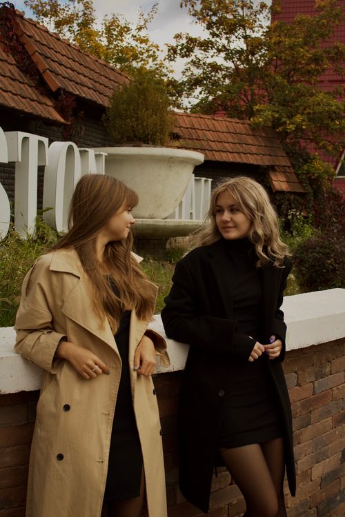 Free Two women in stylish coats sharing a moment outdoors against a rustic backdrop. Stock Photo