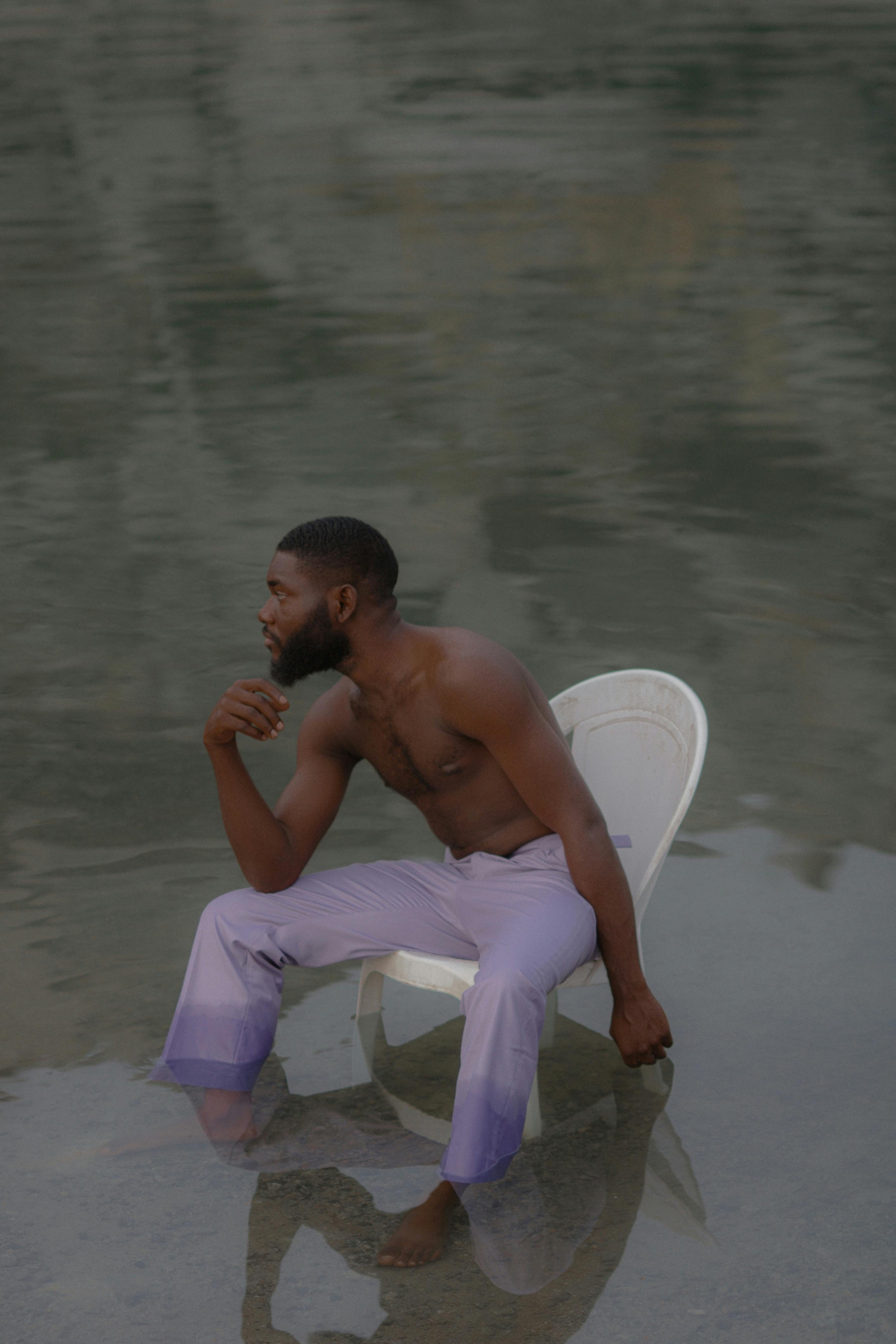 A shirtless man sitting on a chair in shallow water, deep in thought.
