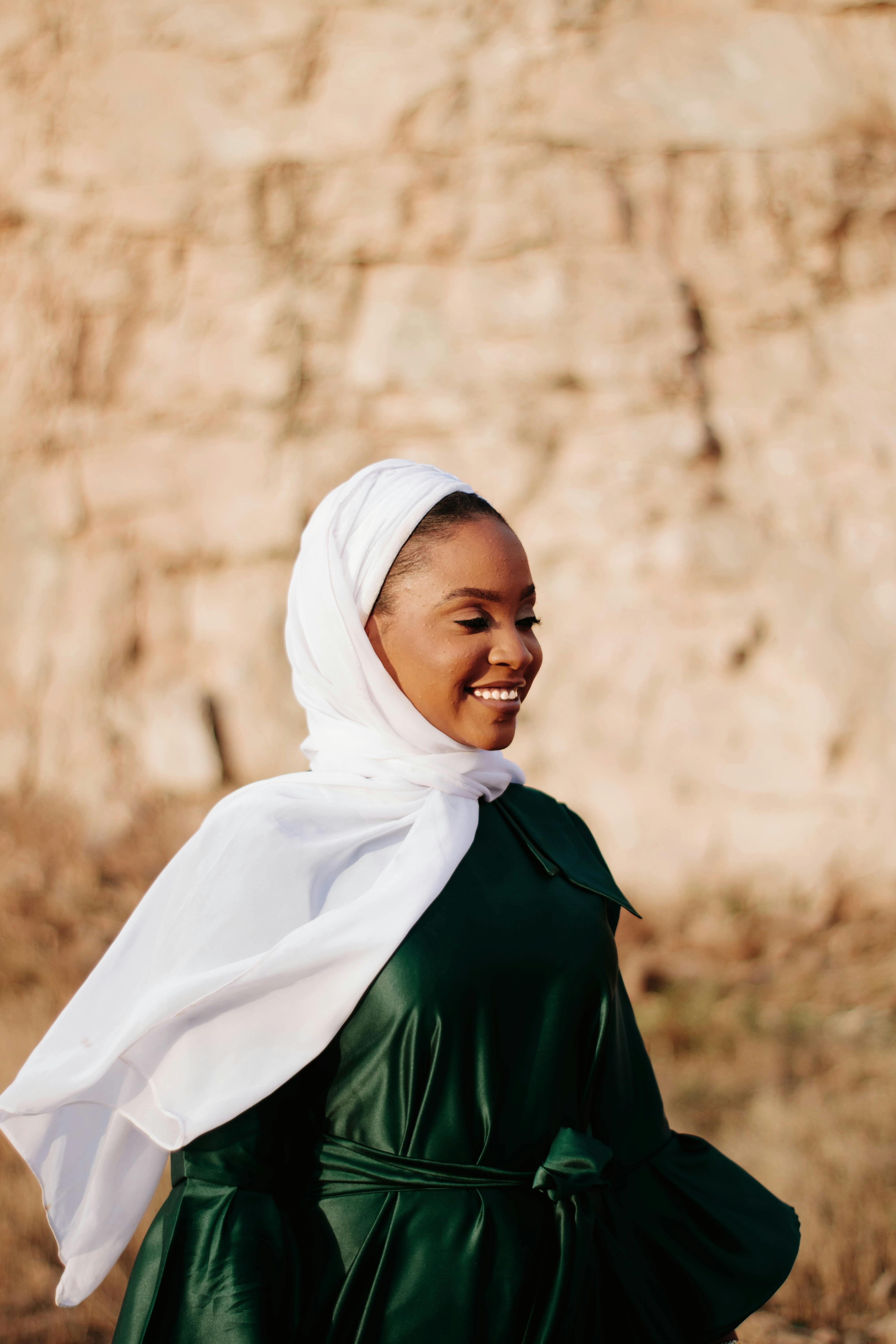 A woman in a green dress and hijab smiling outdoors against a natural background.