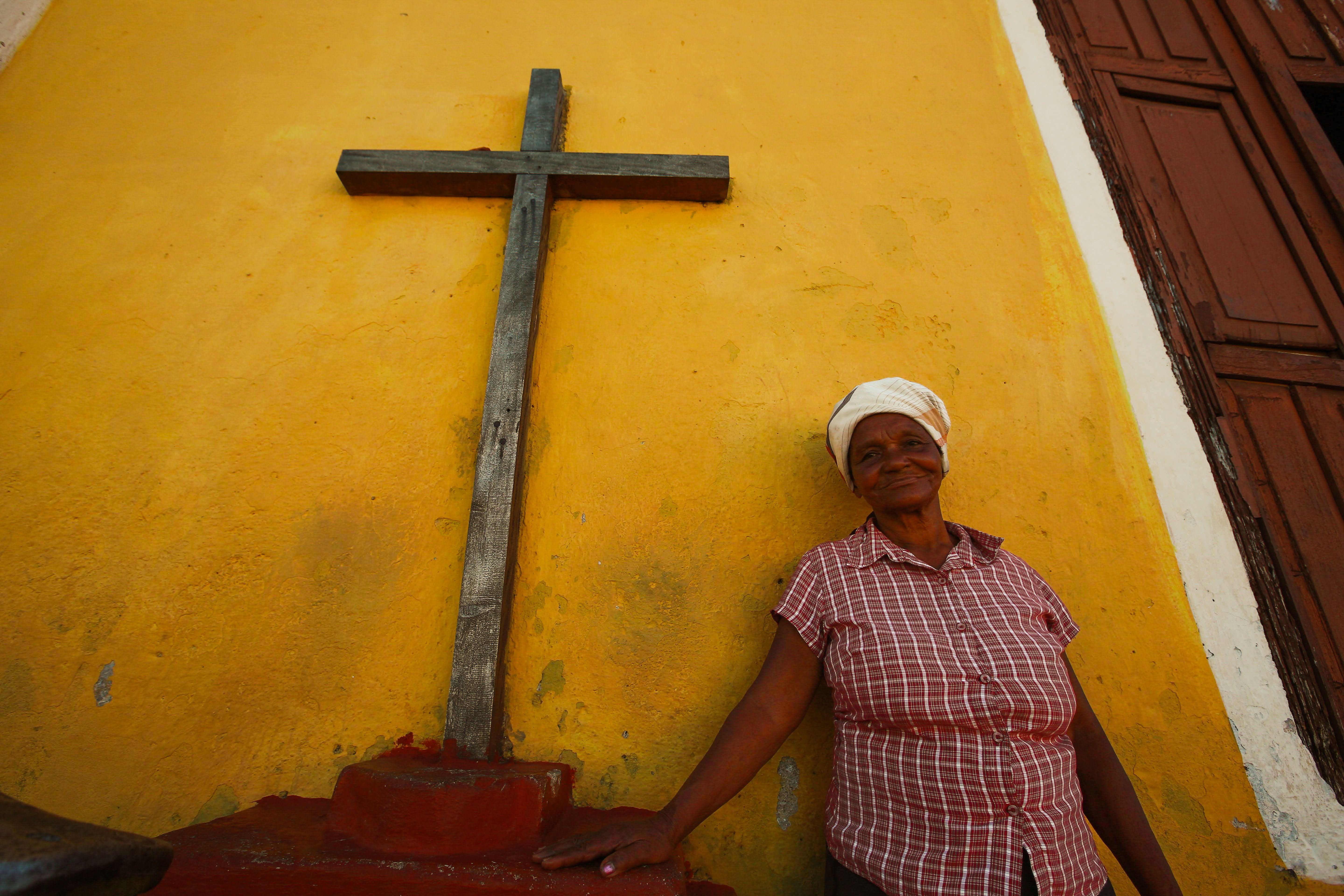 An elderly woman in a plaid shirt stands by a large wooden cross against a vibrant yellow wall.