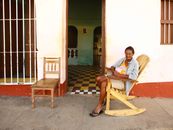A woman sits on a wooden rocking chair on a sunny day outside a rustic building with a tiled floor.