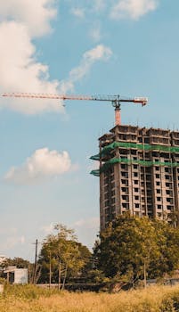 A high-rise building under construction using a tower crane on a sunny day with greenery nearby.