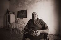Sepia-toned image of an elderly man sitting in a rustic living room, smoking a cigar.